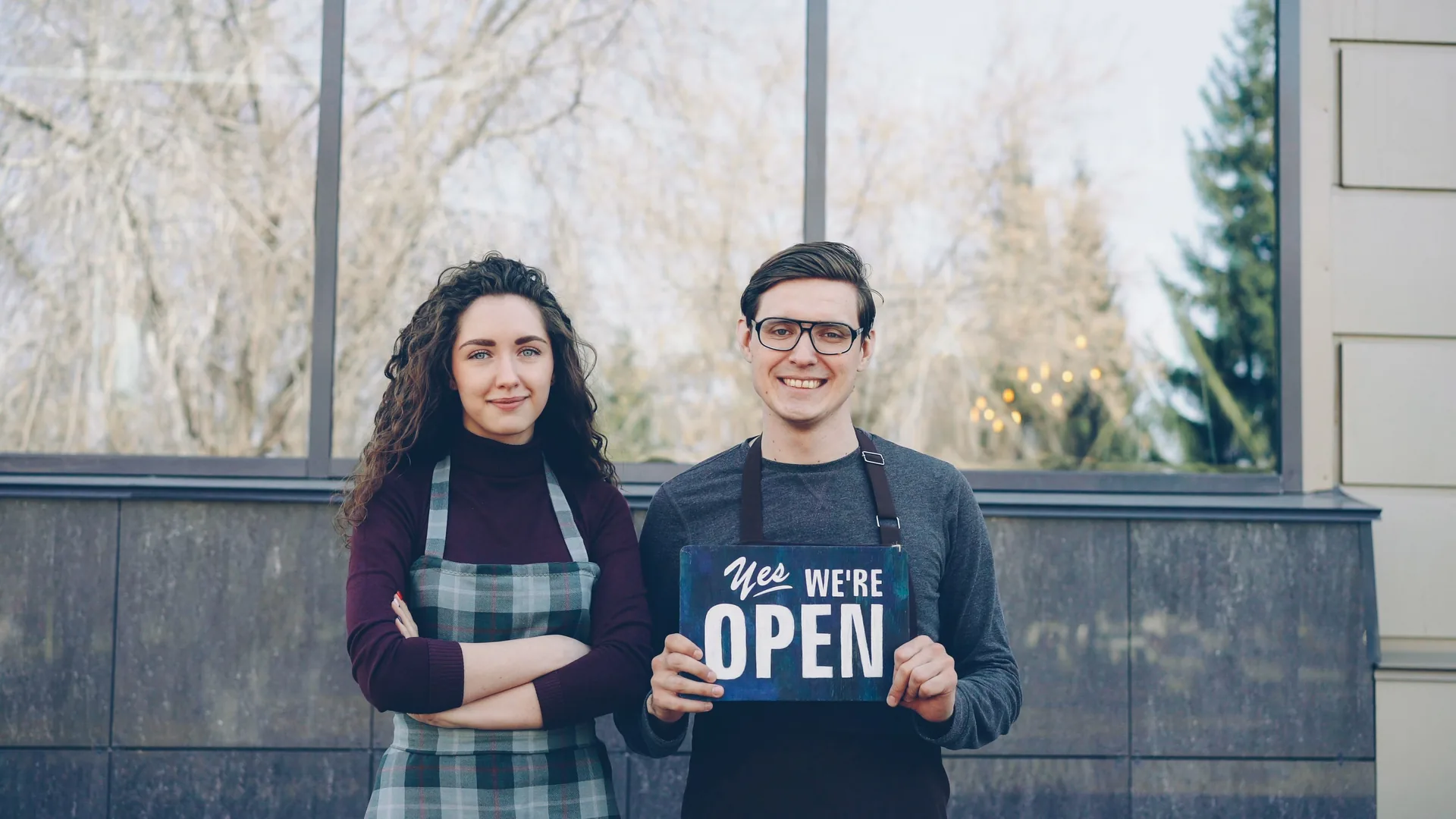 Two business owners holding a Yes We're Open sign outside their shop